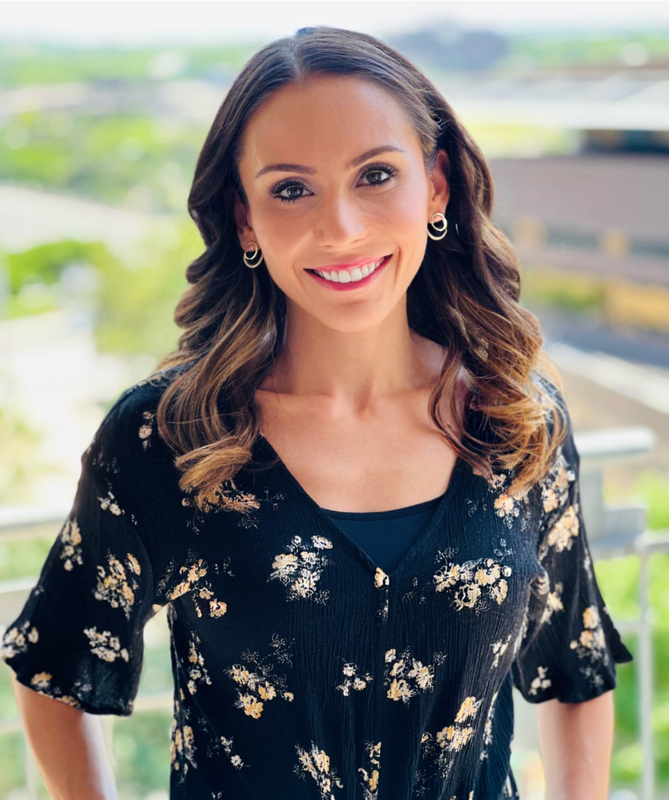 Woman with wavy brown hair and hoop earrings, wearing a black floral top, standing outdoors with a blurred background of greenery and buildings.