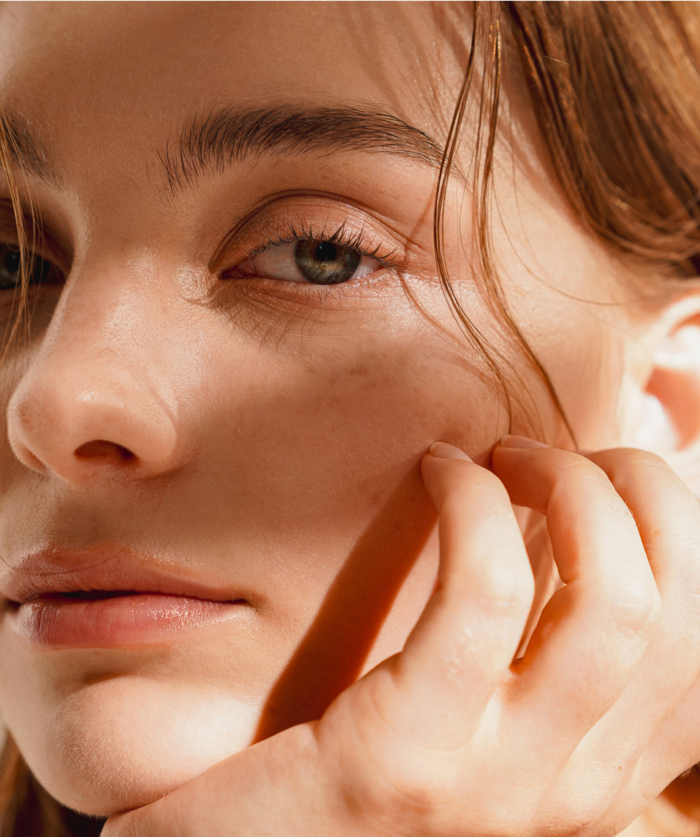 Close-up of a person’s face with light brown hair, touching their cheek, showing natural skin texture and a calm expression.