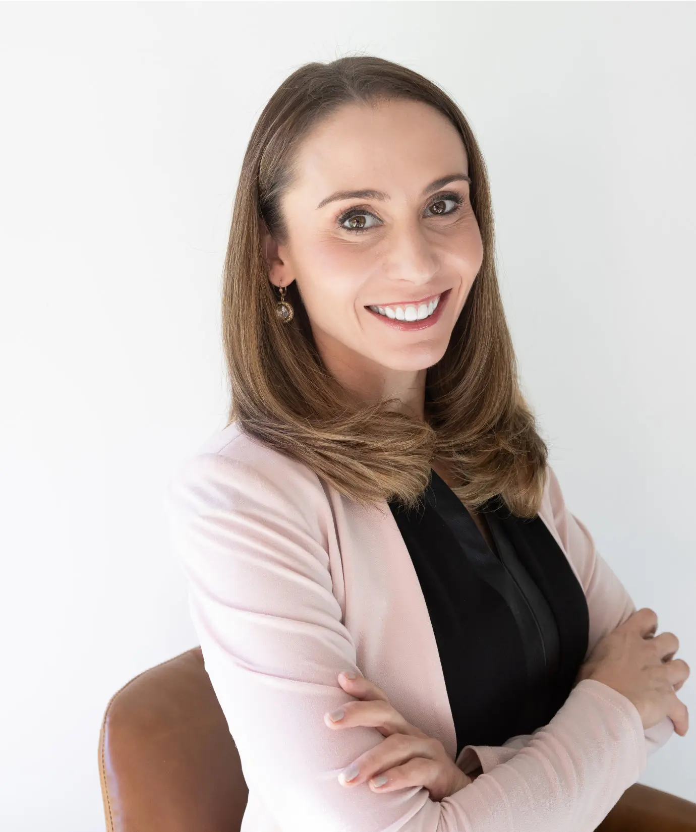 Woman with long brown hair, wearing a pink blazer over a black top, sits on a brown chair with arms crossed, smiling at the camera against a plain white background.