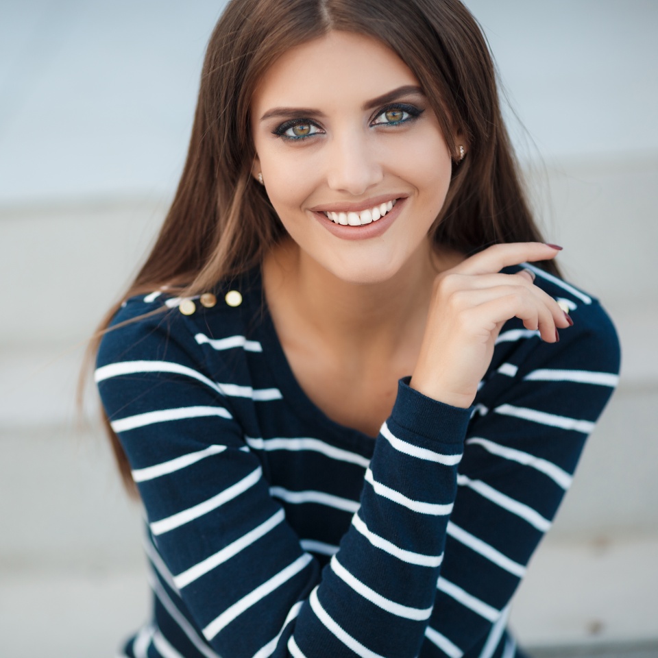 Young beautiful woman brunette with long straight hair and gray-green eyes,a nice smile and white straight teeth,light makeup,dressed in a striped t-shirt and blue jeans,spends time in the city in the spring,sitting on the steps,one in the fresh air