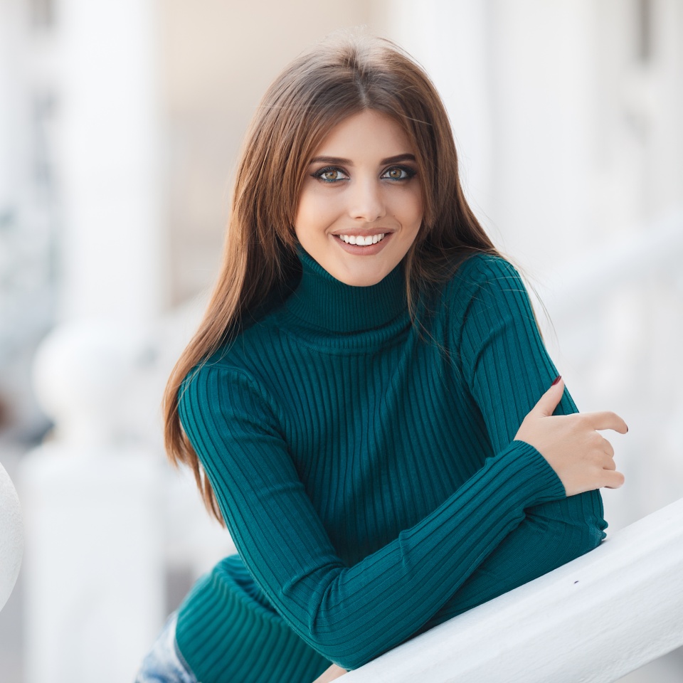 Young beautiful brunette woman with long straight hair and grey eyes,nice smile and white straight teeth,light makeup,dressed in a green sweater and blue jeans,hanging out in the city in the spring,standing on a white terrace,one in the fresh air