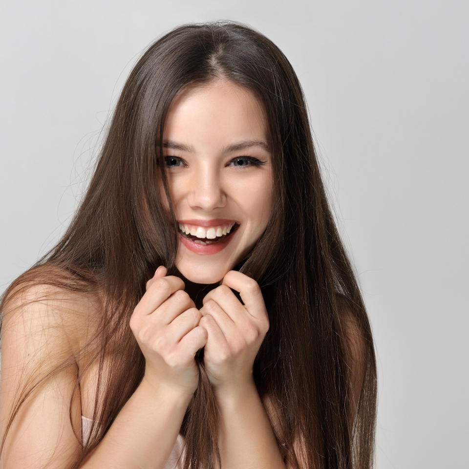 Laughing woman covers her face with long hair. Beautiful woman with bare shoulders has a clean well-groomed skin and long straight hair. Close-up portrait against a light gray background.