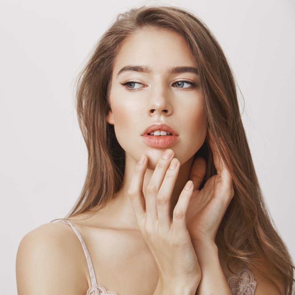 Makeup underline natural beauty. Studio shot of affective romantic caucasian woman with clean skin gently touching chin and looking aside with dreamy expression, standing over gray background