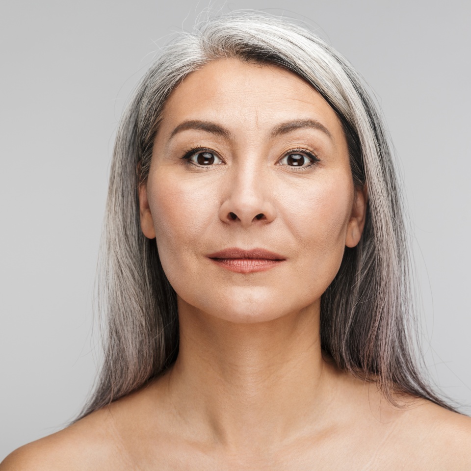 Beauty portrait of an attractive sensual mature topless woman with long gray hair standing isolated over gray background, looking at camera