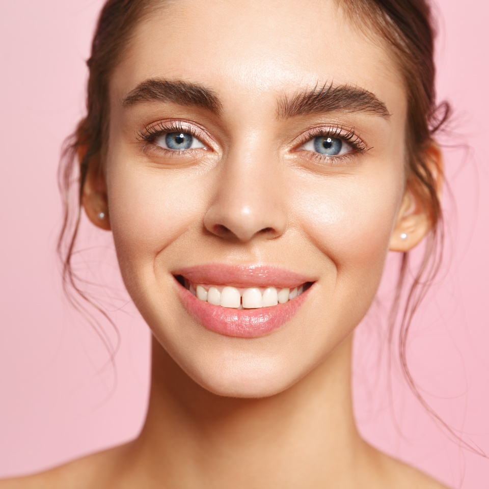 Portrait of caucasian young woman with blue eyes and perfect white smile, nude lips gloss and no makeup natural look, gazing happy at camera with clean hydrated facial skin, pink background.