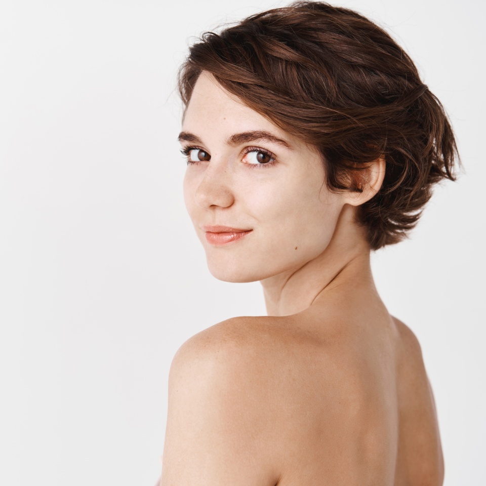 Skincare. Rear view of young caucasian woman turn head back at camera, standing half naked on white background and smiling. Tender girl with no makeup and natural beauty.