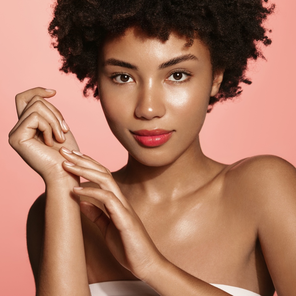 Young woman applying soft skin care cream on her body after shower, gently touching arm with fingertips, standing against pink background, perfect healthy body and skin