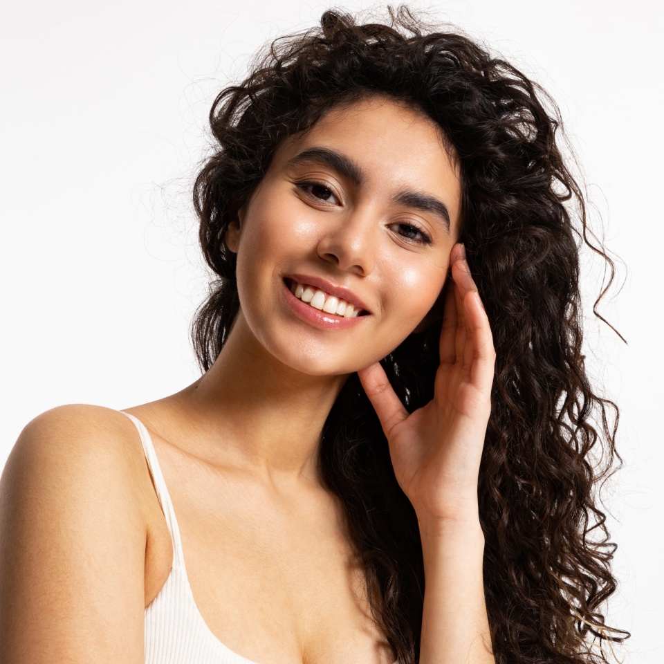 Young woman with thick black and curly hair and glowing skin smiles and looks at the camera.