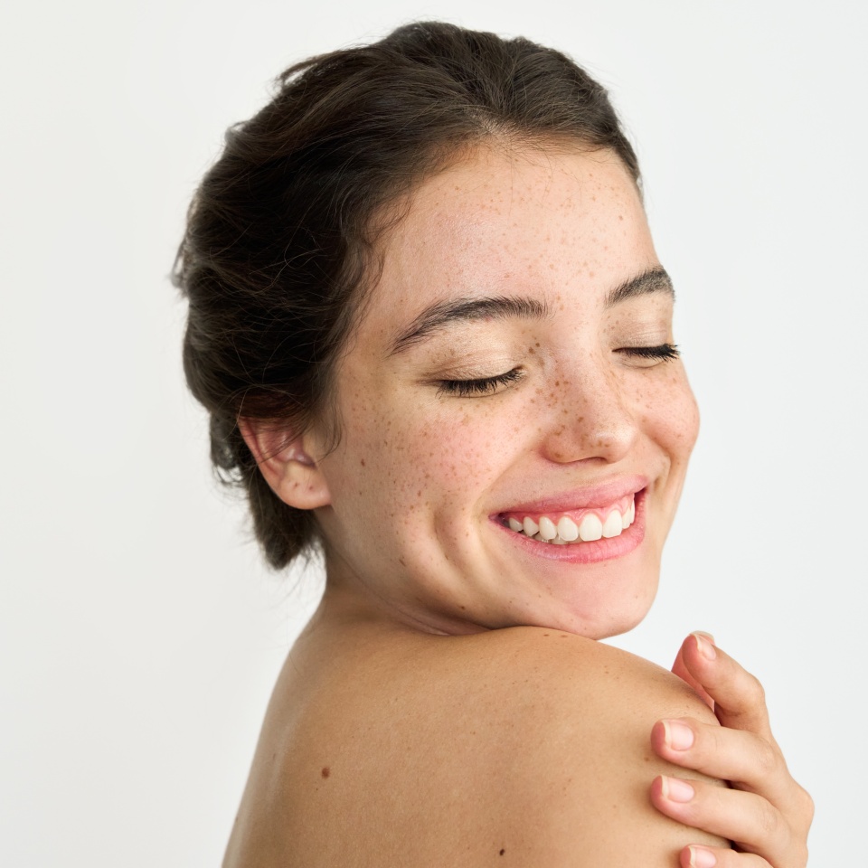 Aesthetic beauty portrait of Latin girl on white background. Young happy brunette woman model with freckles on face touching smooth soft body skin. Skincare cosmetic, skin and body care products ad.