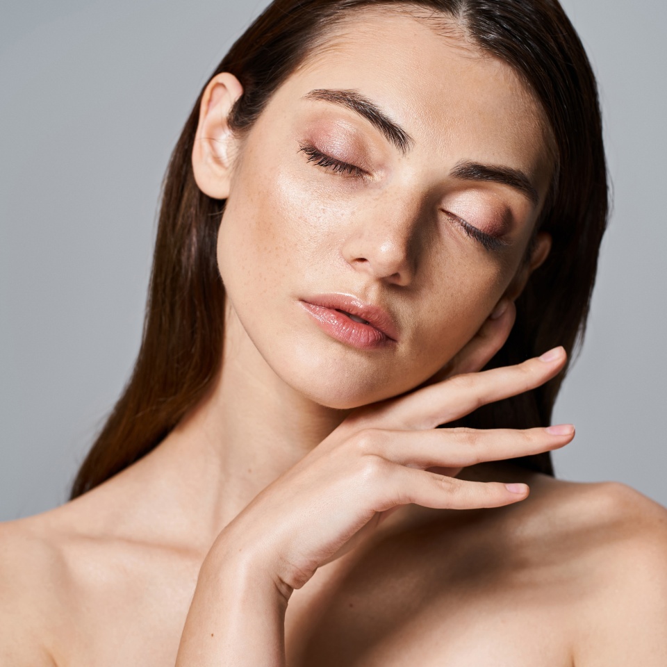 A young, caucasian woman with brunette hair and clean skin posing nude with her eyes gently closed in a studio setting.