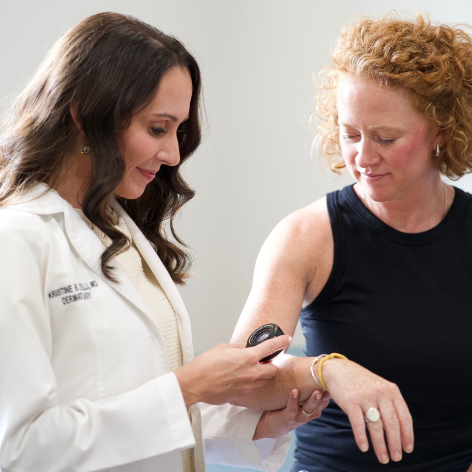A doctor examines a patient’s arm using a handheld device, while the patient looks on and holds her arm out for inspection.