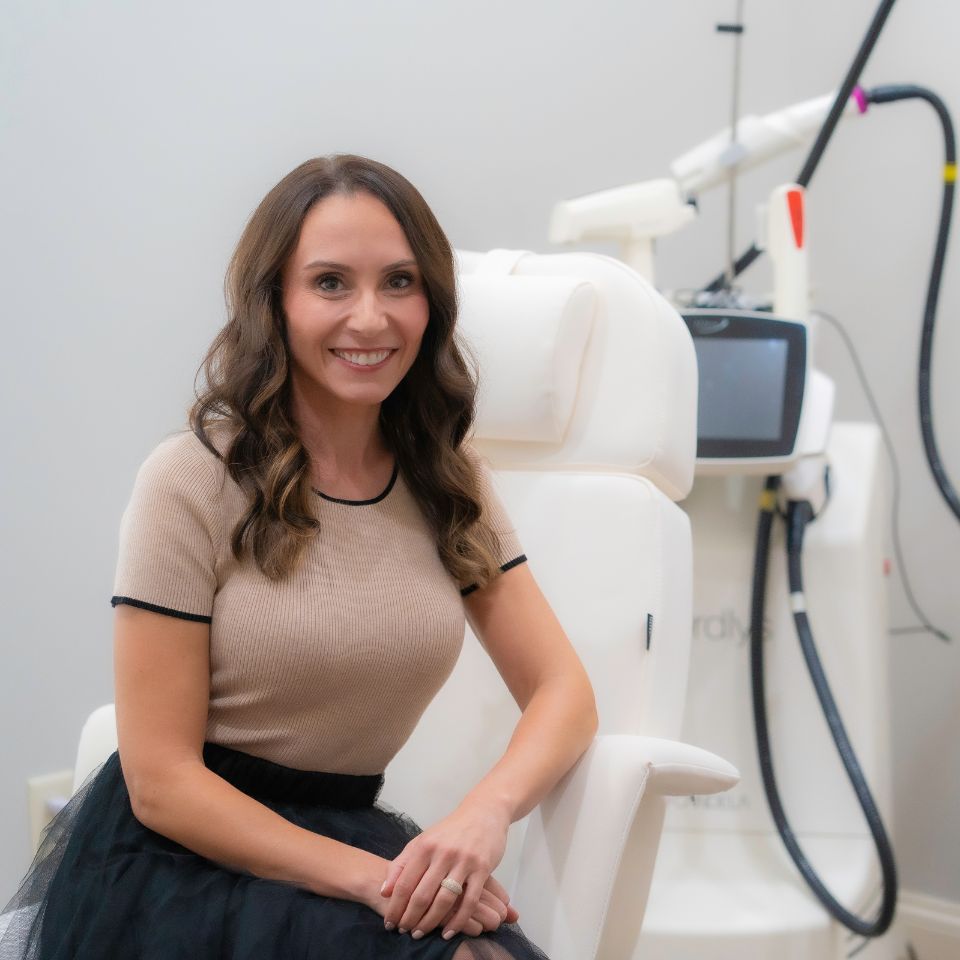 A woman with long brown hair sits on a white chair in a medical or aesthetic clinic, smiling, with a large machine and monitor in the background.