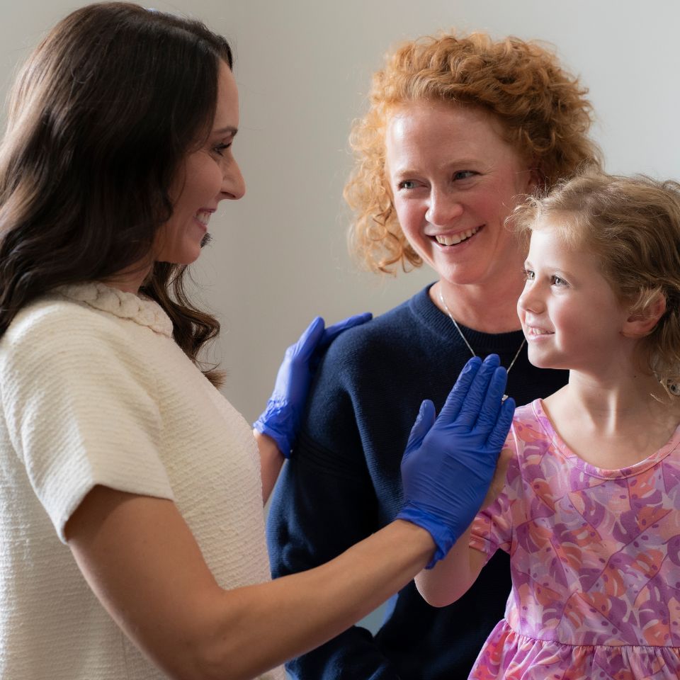 A woman wearing blue medical gloves smiles and high-fives a young girl in a pink dress, while another woman stands beside them, smiling.