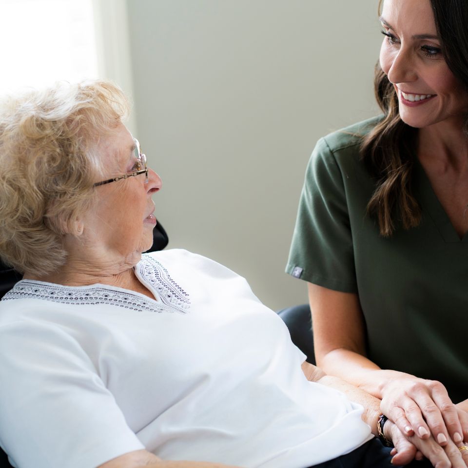 An elderly woman sits in a chair while a caregiver in green scrubs sits beside her, holding her hand and smiling.