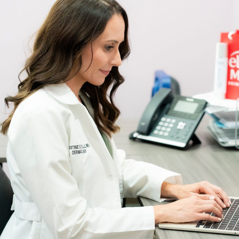 A woman in a white lab coat sits at a desk typing on a laptop, with an office phone and supplies in the background.