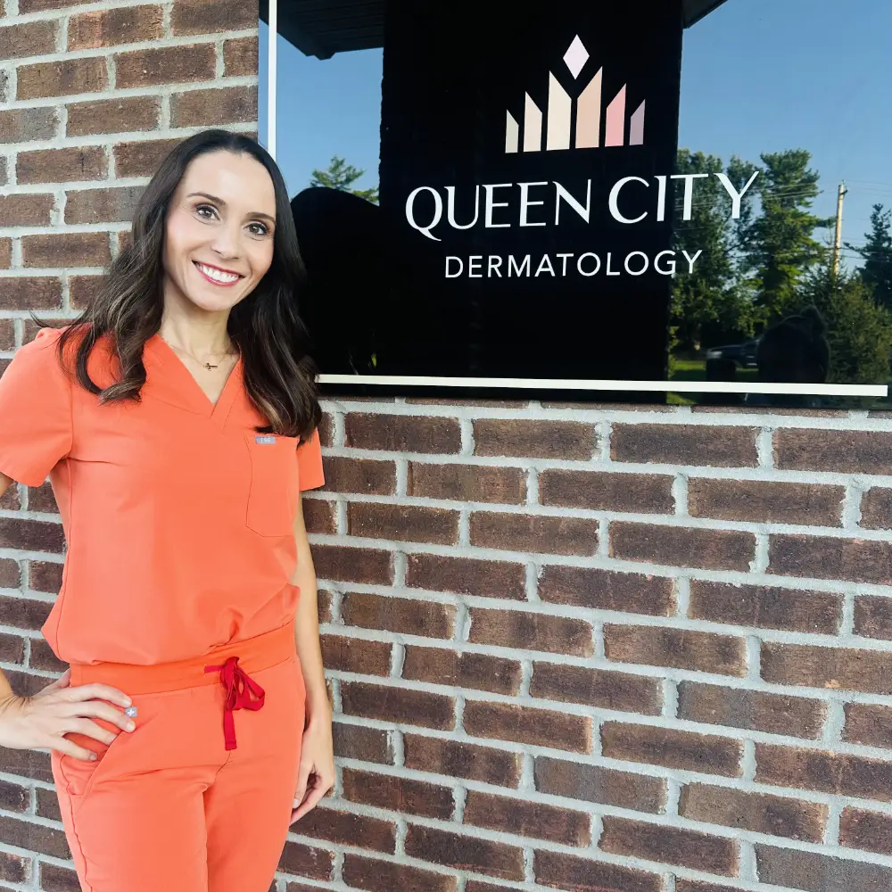 Dr. Kristine Zitelli in coral scrubs posing outside Queen City Dermatology clinic next to the practice sign on a brick building.