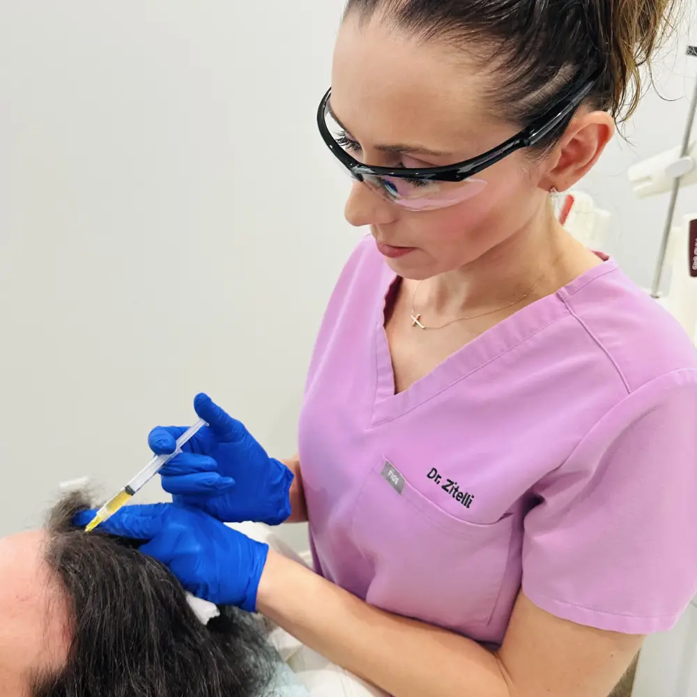Dr. Kristine Zitelli in pink scrubs and protective eyewear administering an injection to a patient's scalp area.