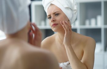 Young woman sits in the bathroom in front of the makeup mirror and does cosmetic procedures. Beautiful girl in white towel. Skin care, health, rejuvenation and spa treatment concept.